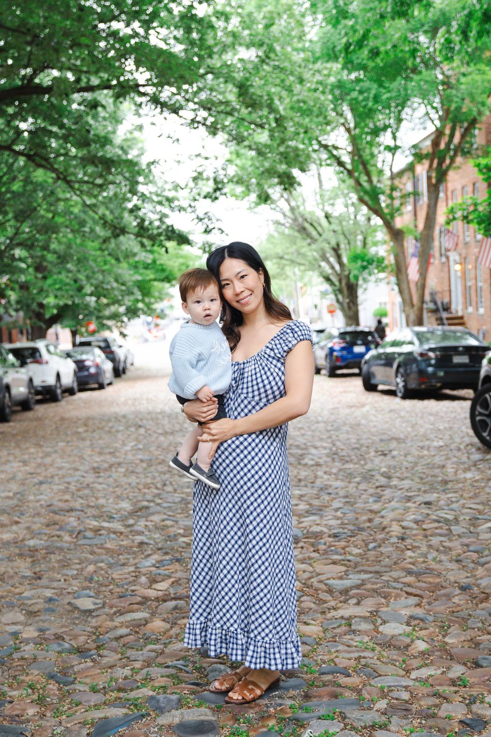 A smiling Asian woman in a blue gingham dress holds a baby on a charming cobblestone street lined with trees and parked cars.
