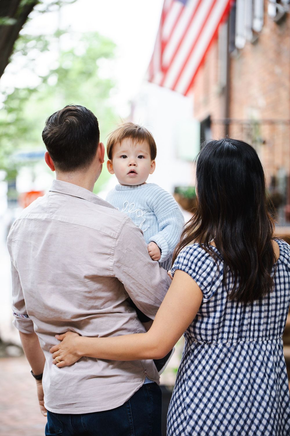 A young family with their baby boy stands on a brick sidewalk near an American flag display.