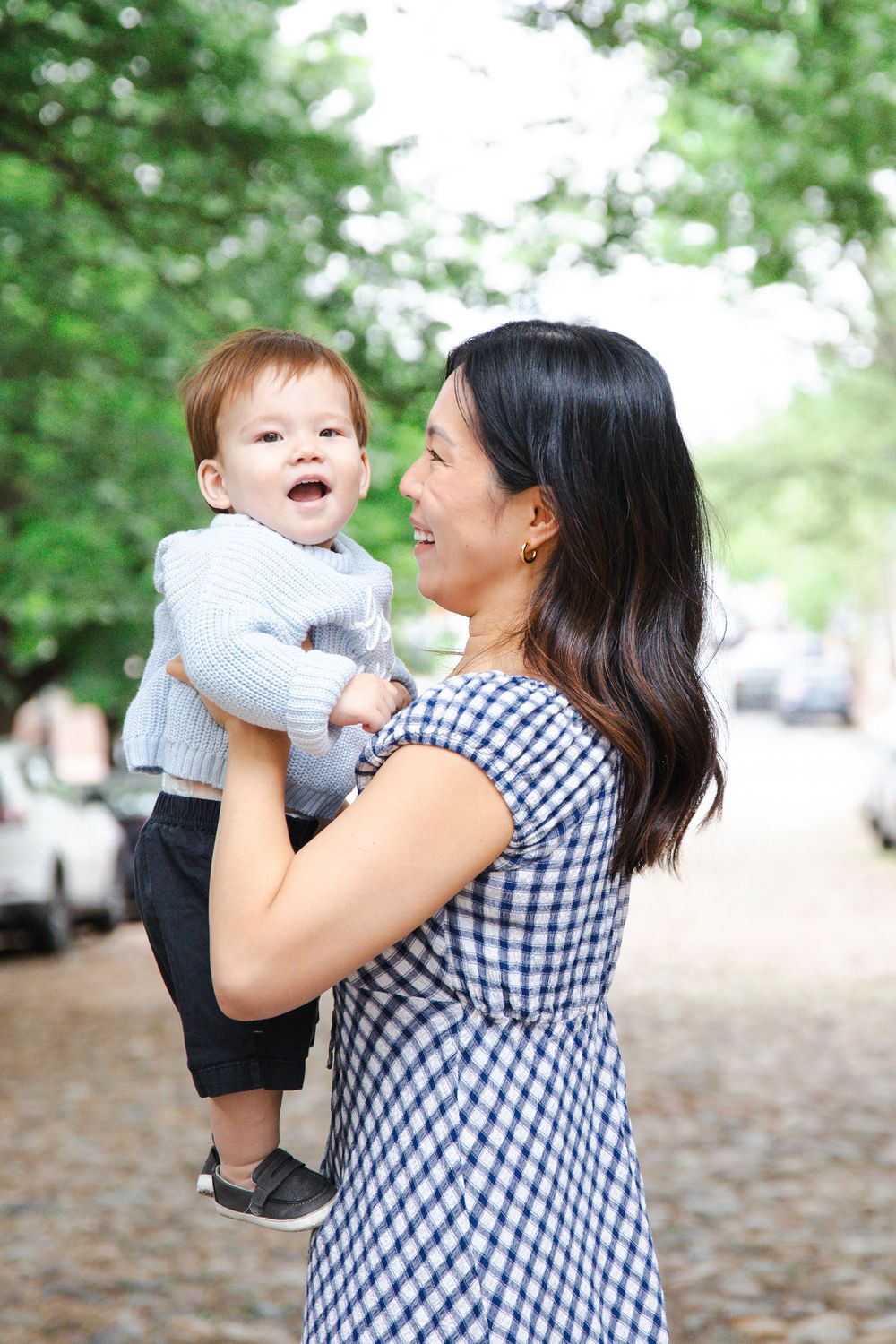 A smiling Asian mother in a gingham dress lifts her happy baby boy outdoors on a tree-lined cobblestone street.