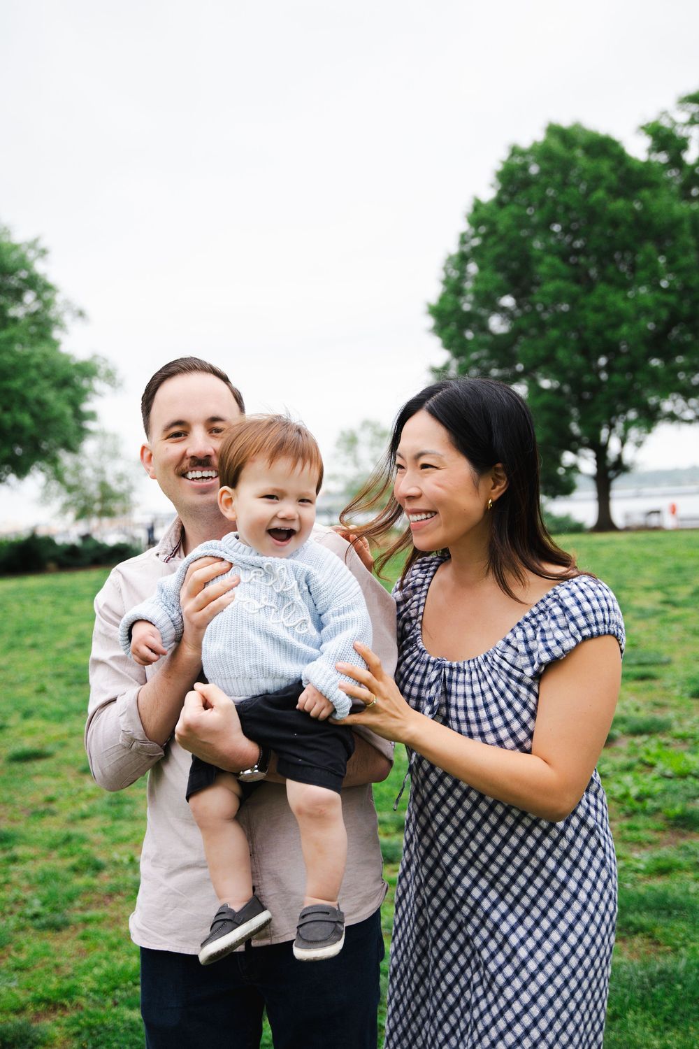 A joyful couple laughs with their giggling baby boy in a green park on an overcast day.