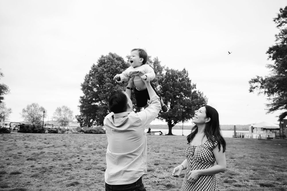 Black and white photo of parents lifting baby in air outdoors in a park setting.