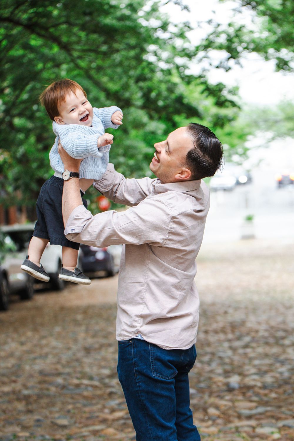 A joyful father lifts his laughing toddler high in the air outdoors on a cobblestone street lined with green trees.