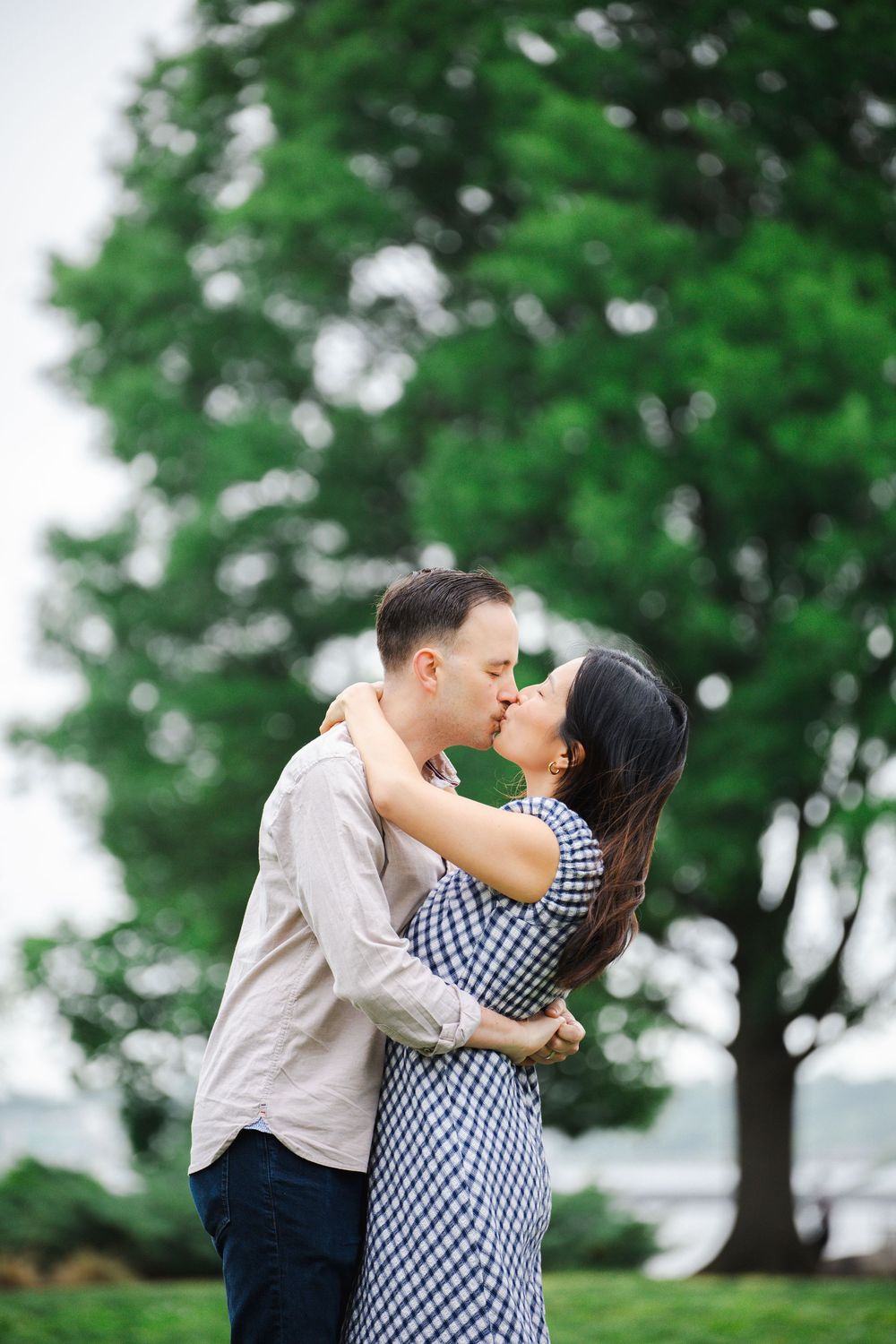 A couple shares a romantic kiss in a lush green park, the woman in a blue gingham dress embraced by her partner.