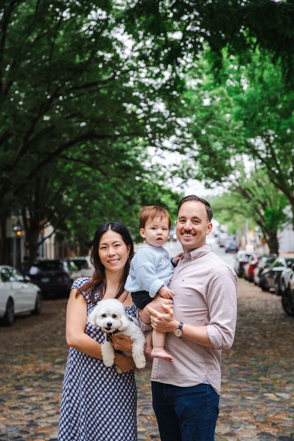 Happy family with baby and white dog posing on cobblestone street lined with green trees.