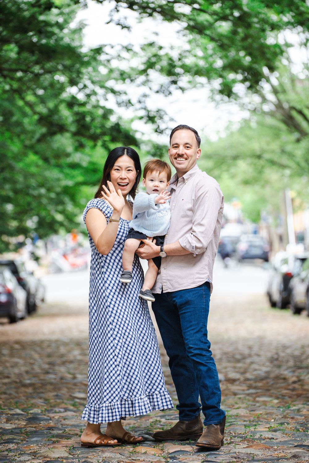 A happy family of three poses on a cobblestone street, with a woman in a blue gingham dress and a man holding their baby.