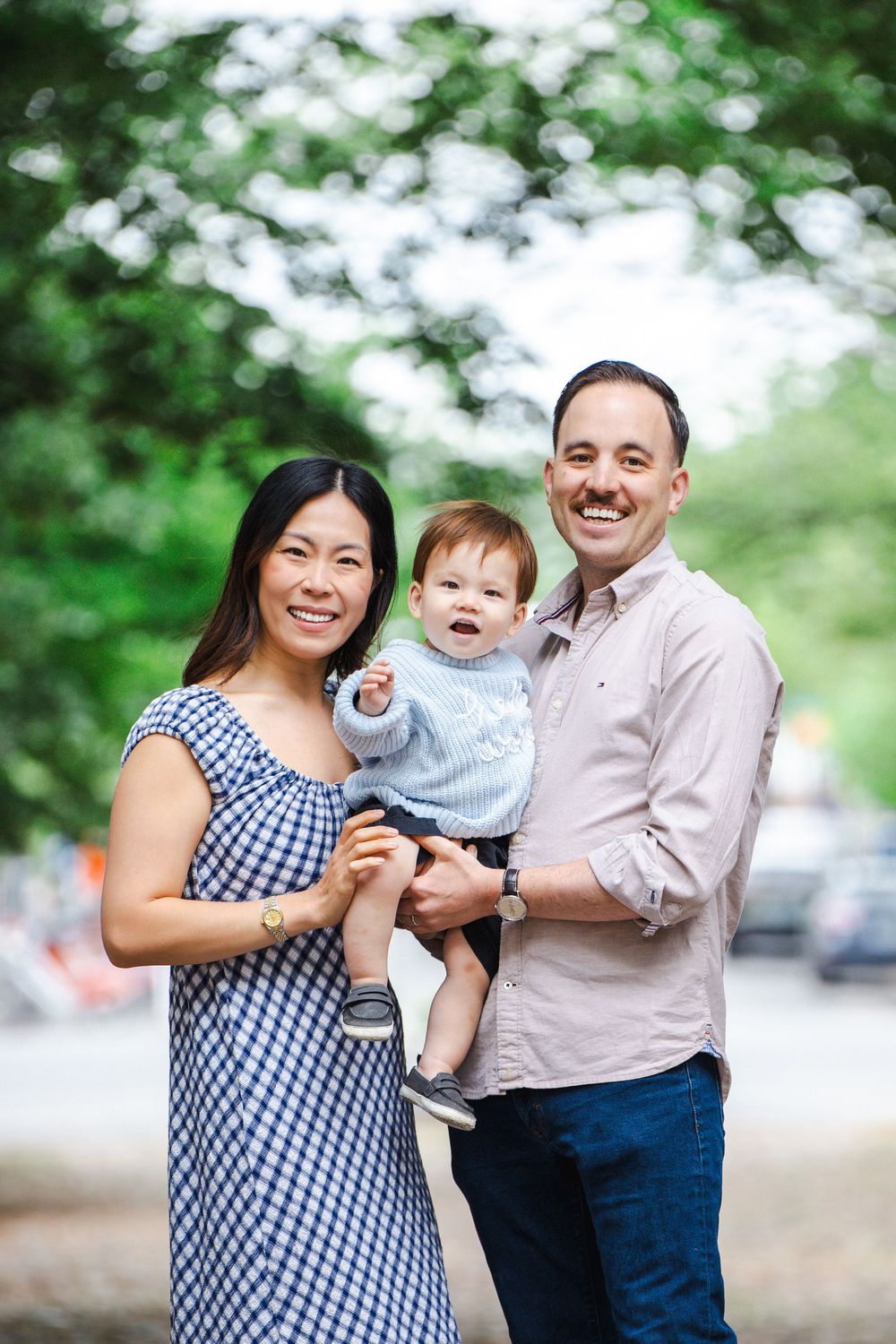 Happy young family with baby boy posing outdoors in park with green trees in background.