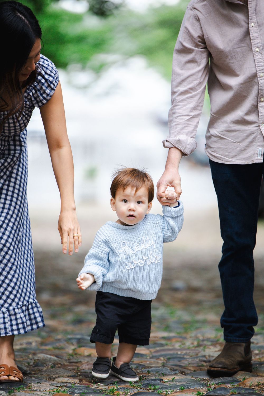 Toddler in blue sweater takes first steps holding parents hands on cobblestone path outdoors.