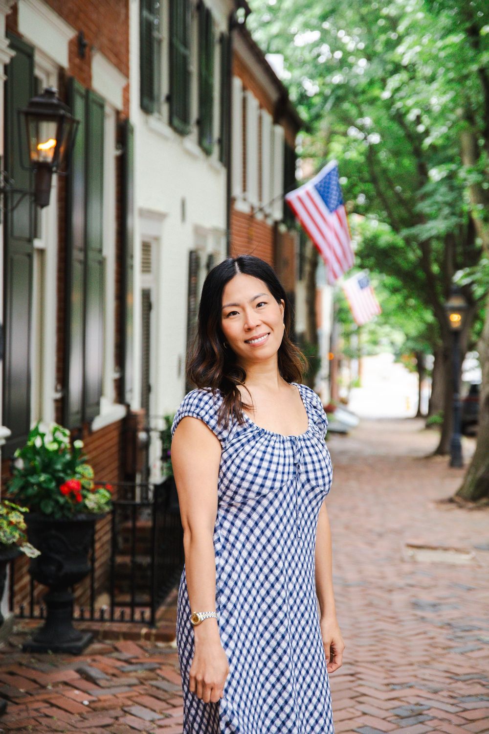 Woman in blue gingham dress smiling on brick sidewalk with American flag and townhouses in background.