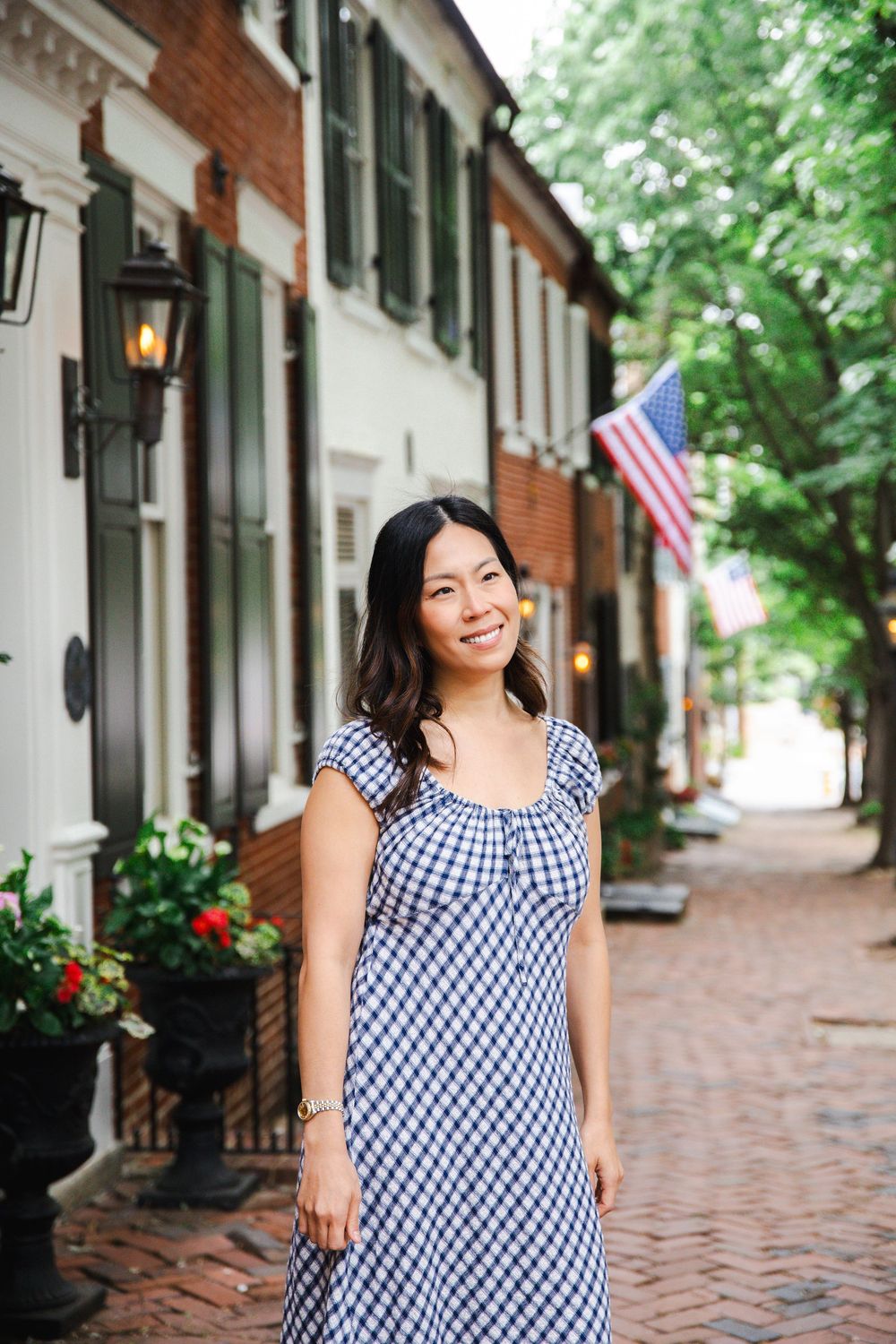 Woman in blue gingham dress smiling on a brick sidewalk with an American flag and historic row houses behind her.