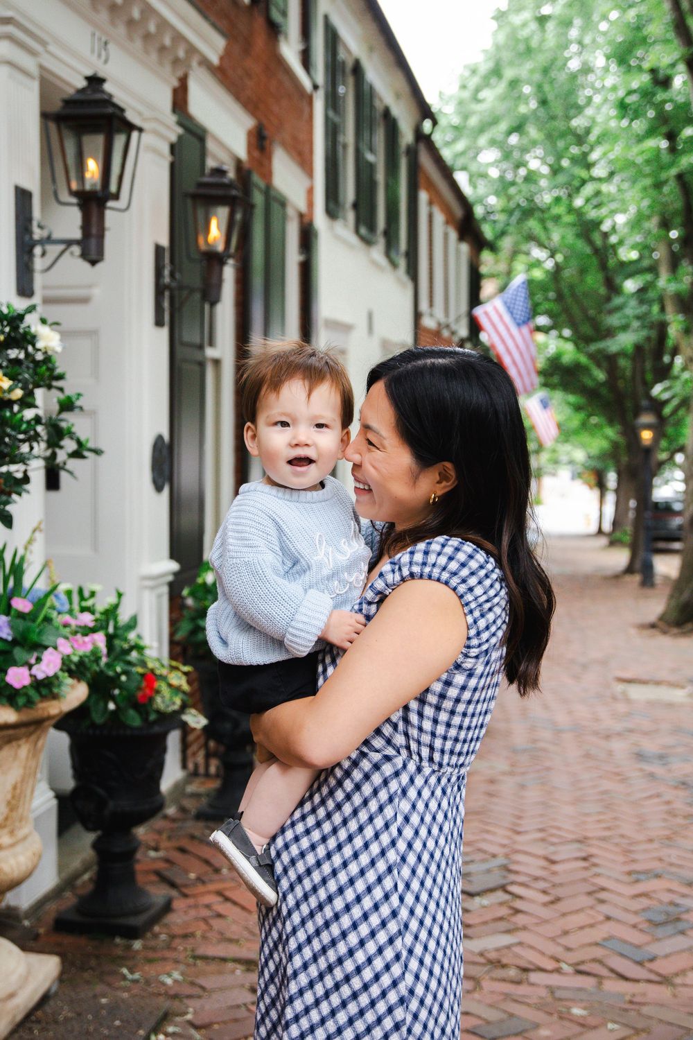 An Asian mother in a gingham dress holds her smiling baby on a charming brick sidewalk with historic townhouses.