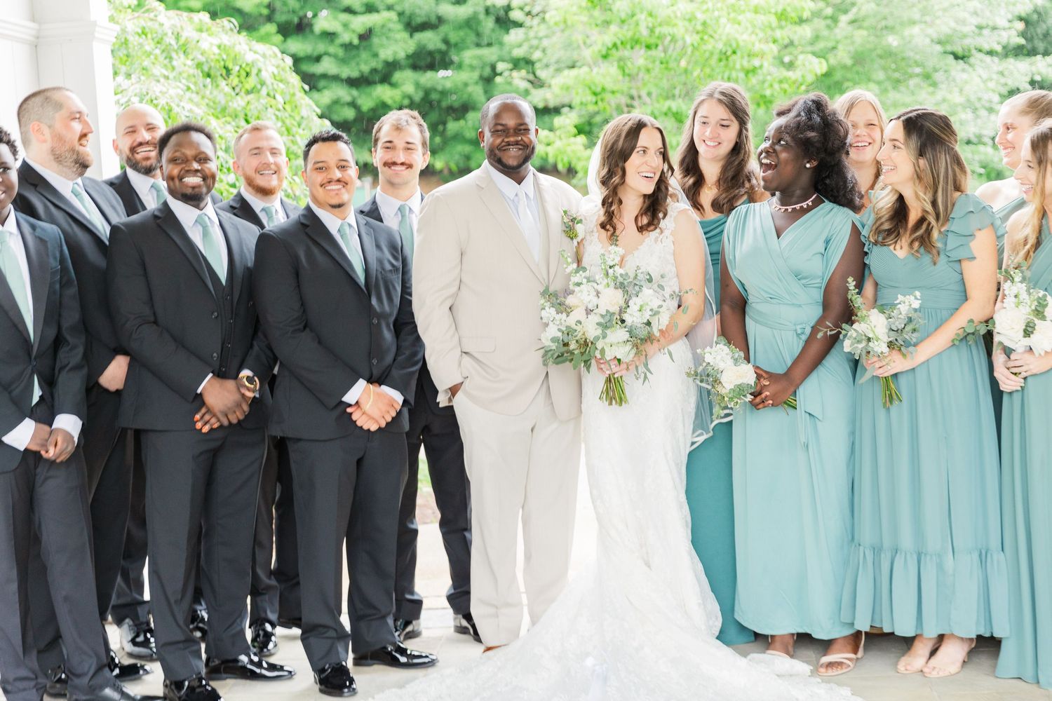 Wedding party poses outdoors, groomsmen in gray suits, bridesmaids in teal dresses, bride in white gown holding bouquet.