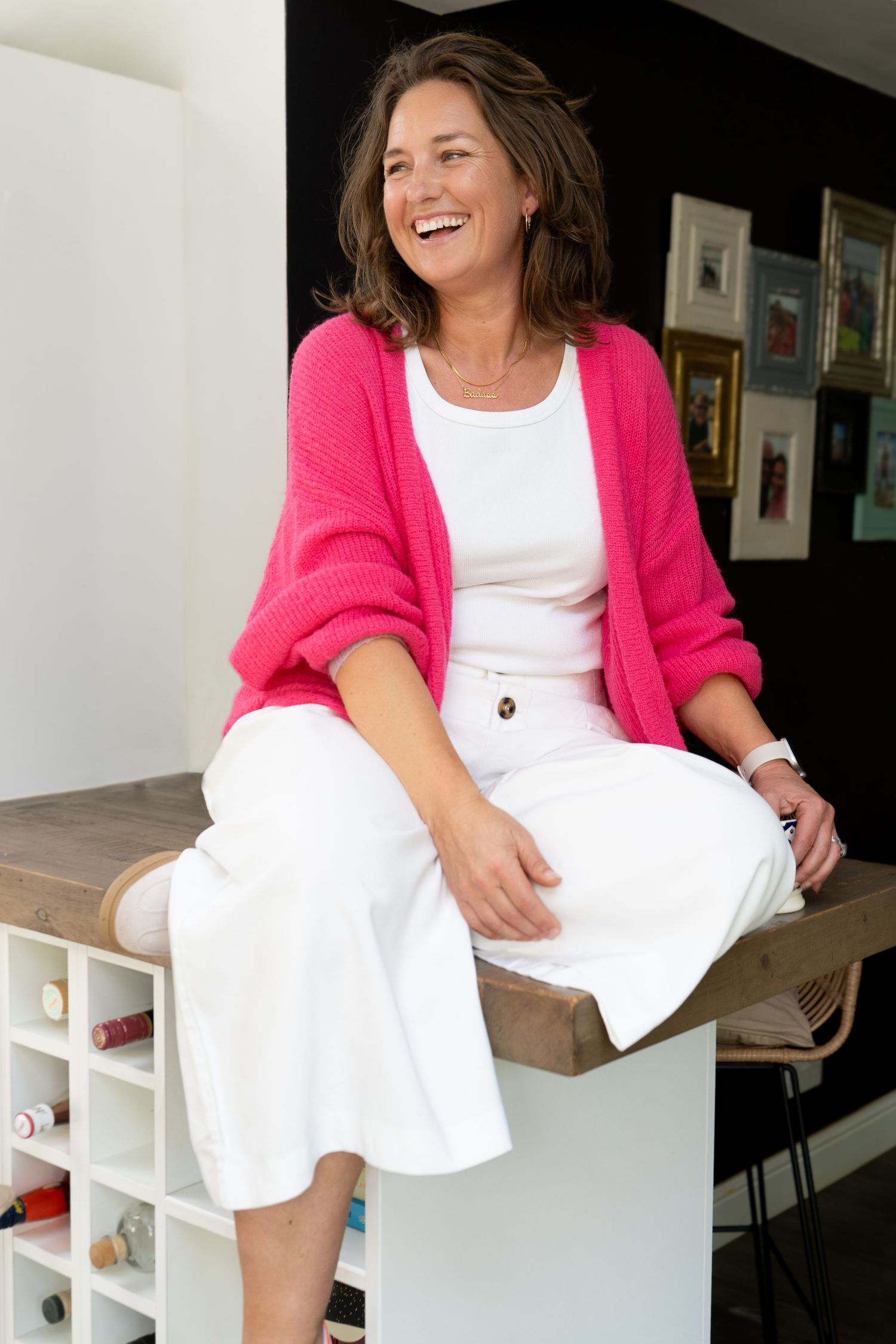 Fun, personal brand photo of female coach in pink cardigan and white outfit sitting on a wooden counter, laughing joyfully in a modern home.