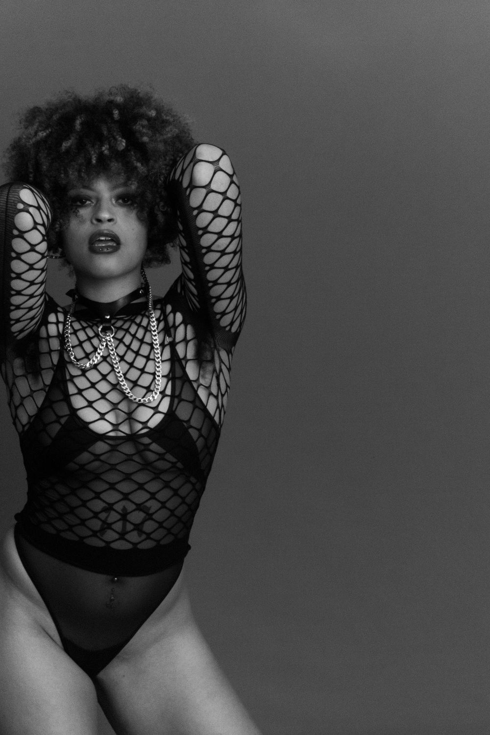 Black and white photo of woman in fishnet bodysuit and chains posing with hands behind head.