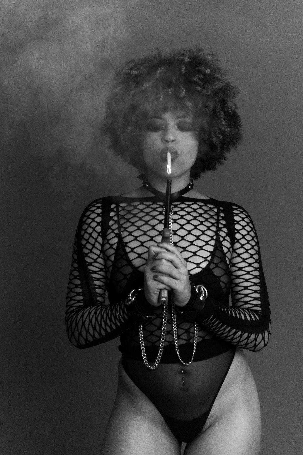 Black and white image of woman in fishnet bodysuit holding cigarette with smoke rising above.
