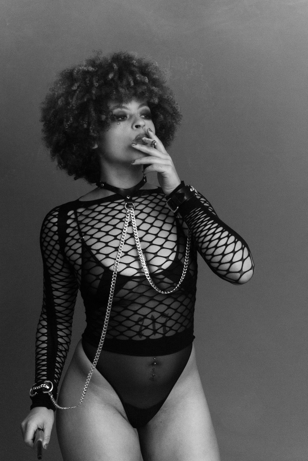 Black and white photo of a woman in fishnet bodysuit with chains posing confidently.