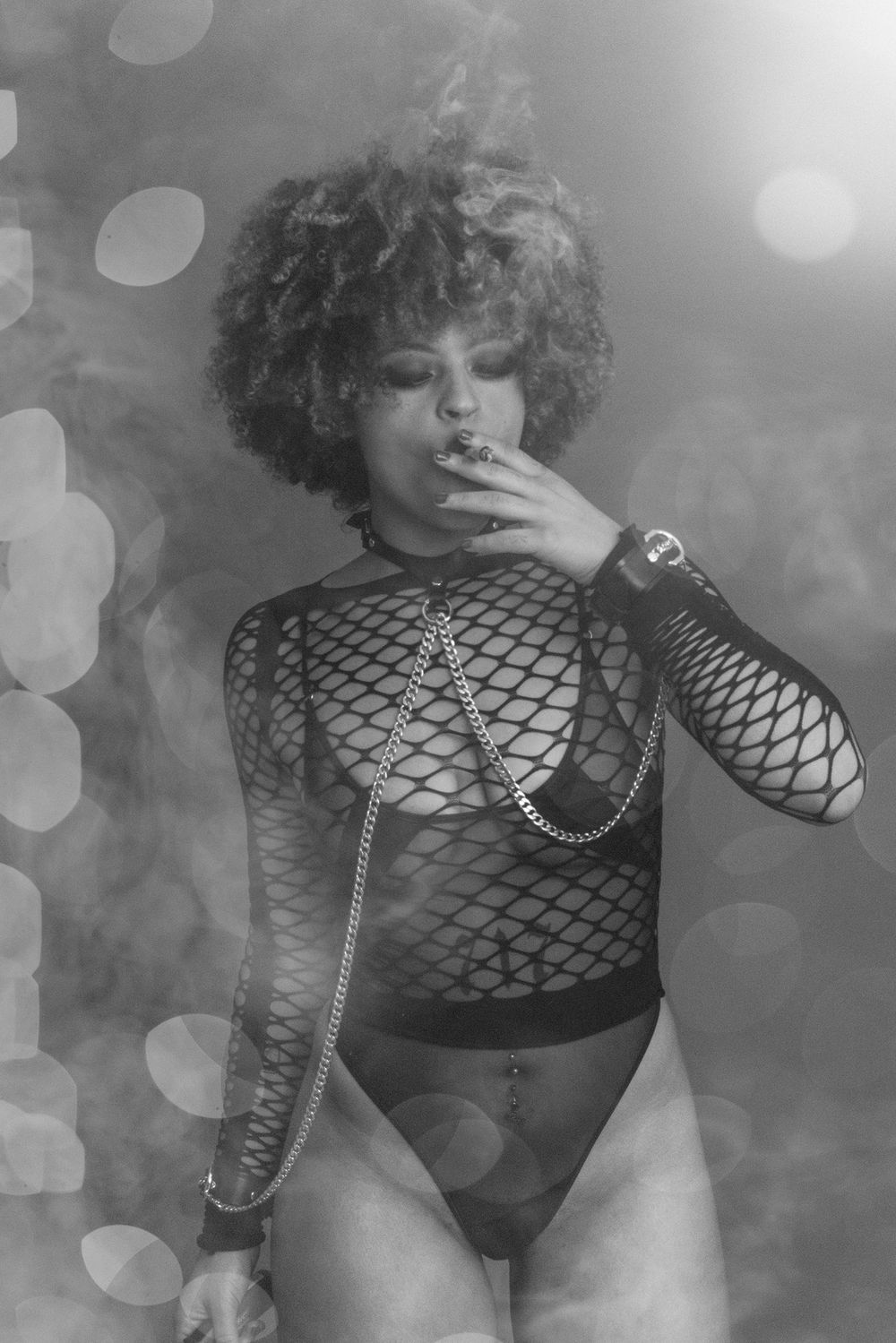 Black and white bokeh photo of woman in fishnet bodysuit smoking with chain accessories.