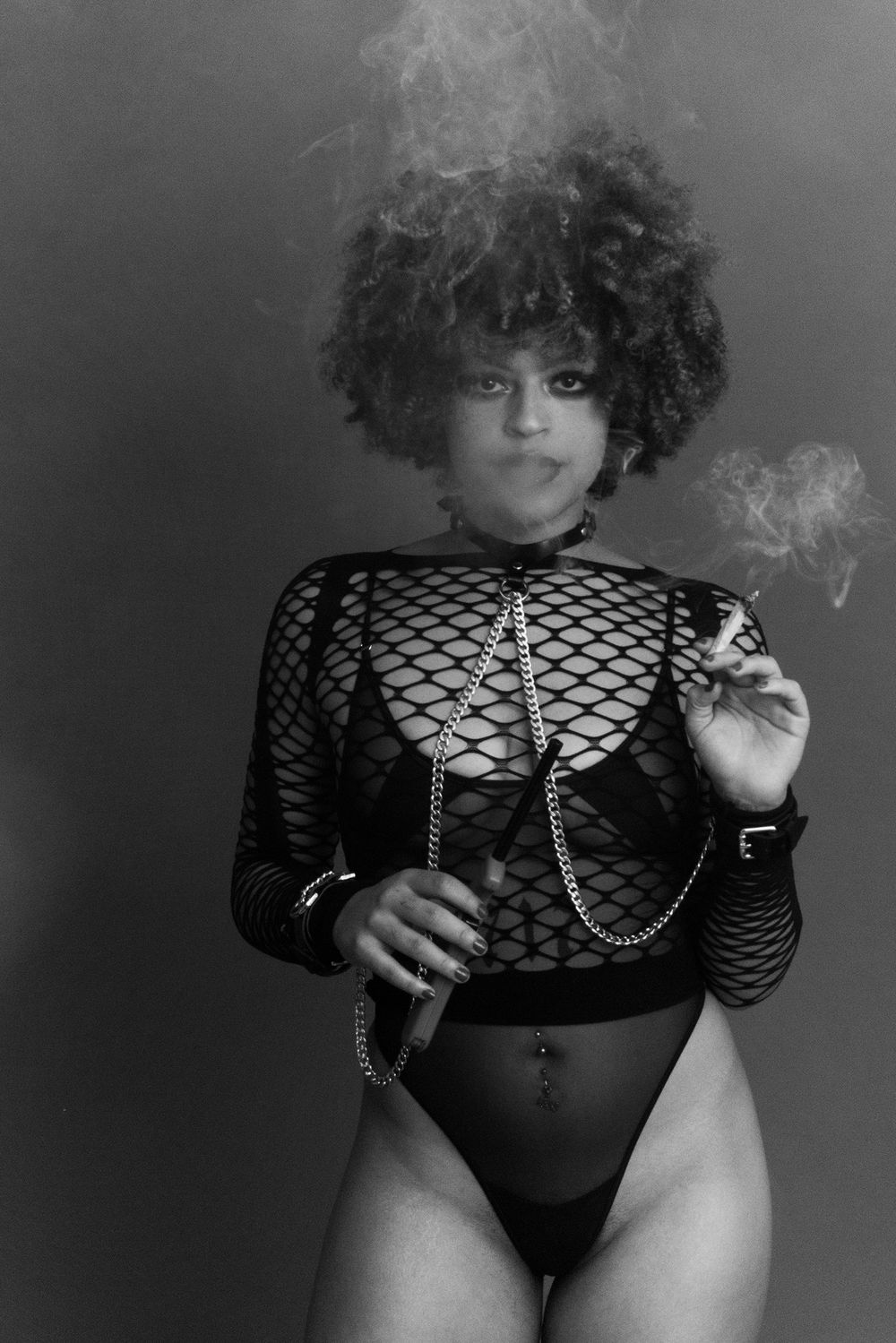 Black and white image of a woman in fishnet bodysuit smoking a cigar with chains.