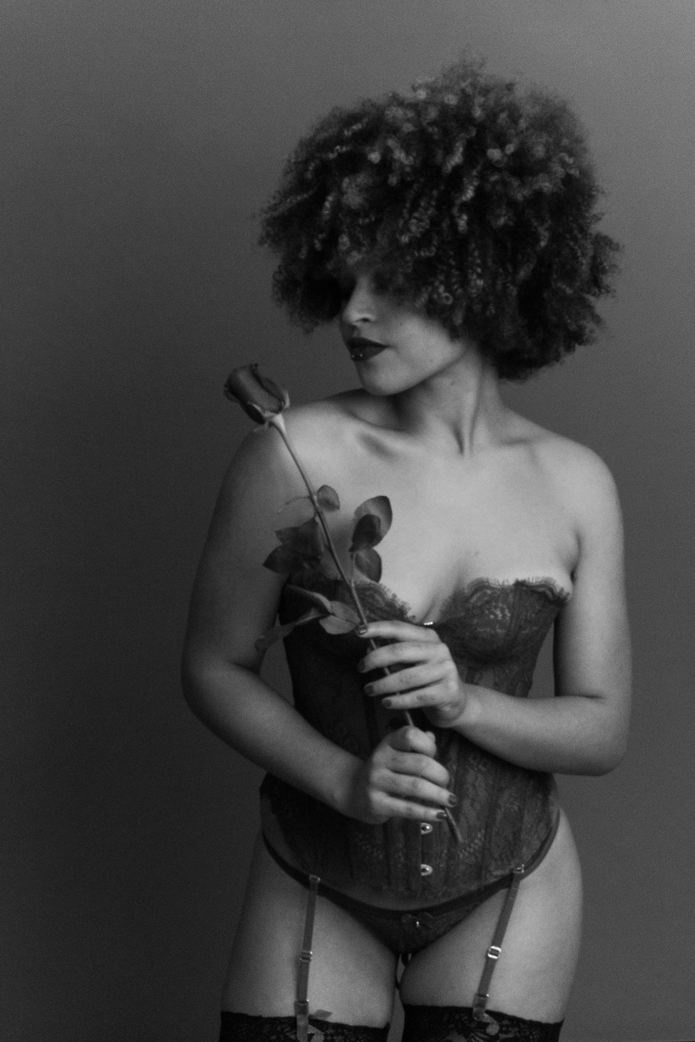 Monochrome portrait of a curly-haired woman in lace corset holding a rose downward.