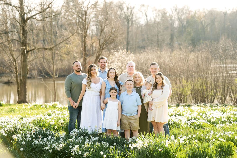 Large extended family group portrait in a blooming daffodil field with a pond and trees in the background.