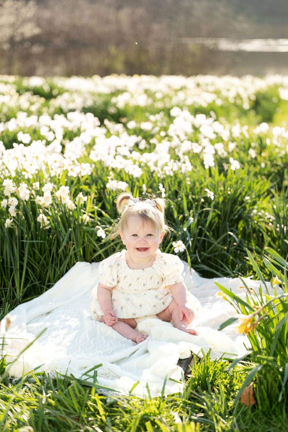 Happy toddler girl in cream romper sitting on blanket surrounded by white daffodils outdoors.