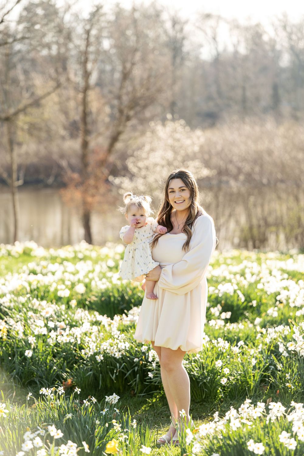 Mother in white dress holding baby girl in floral outfit in blooming daffodil field.