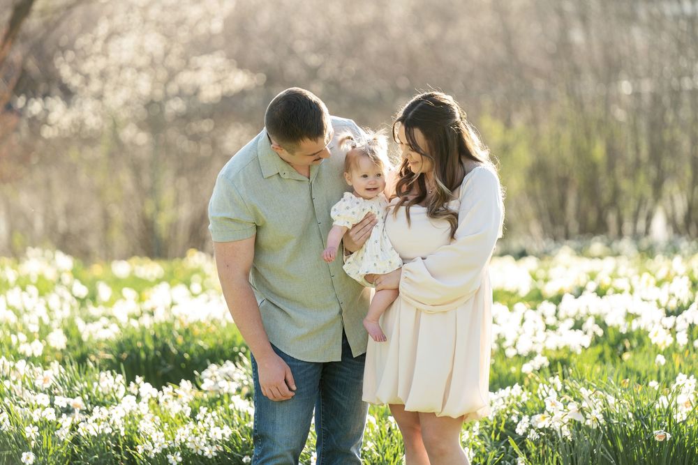 Parents smiling at baby daughter while standing in bright spring daffodil flower field.