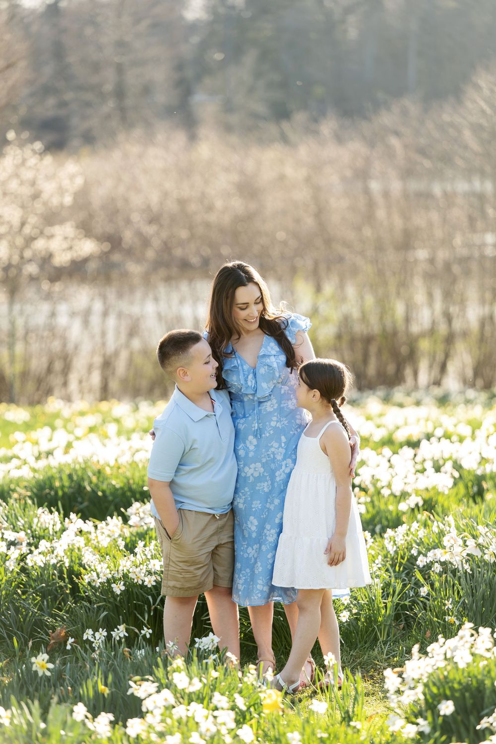 Mother in blue floral dress embraces son and daughter in sunny spring daffodil field portrait.
