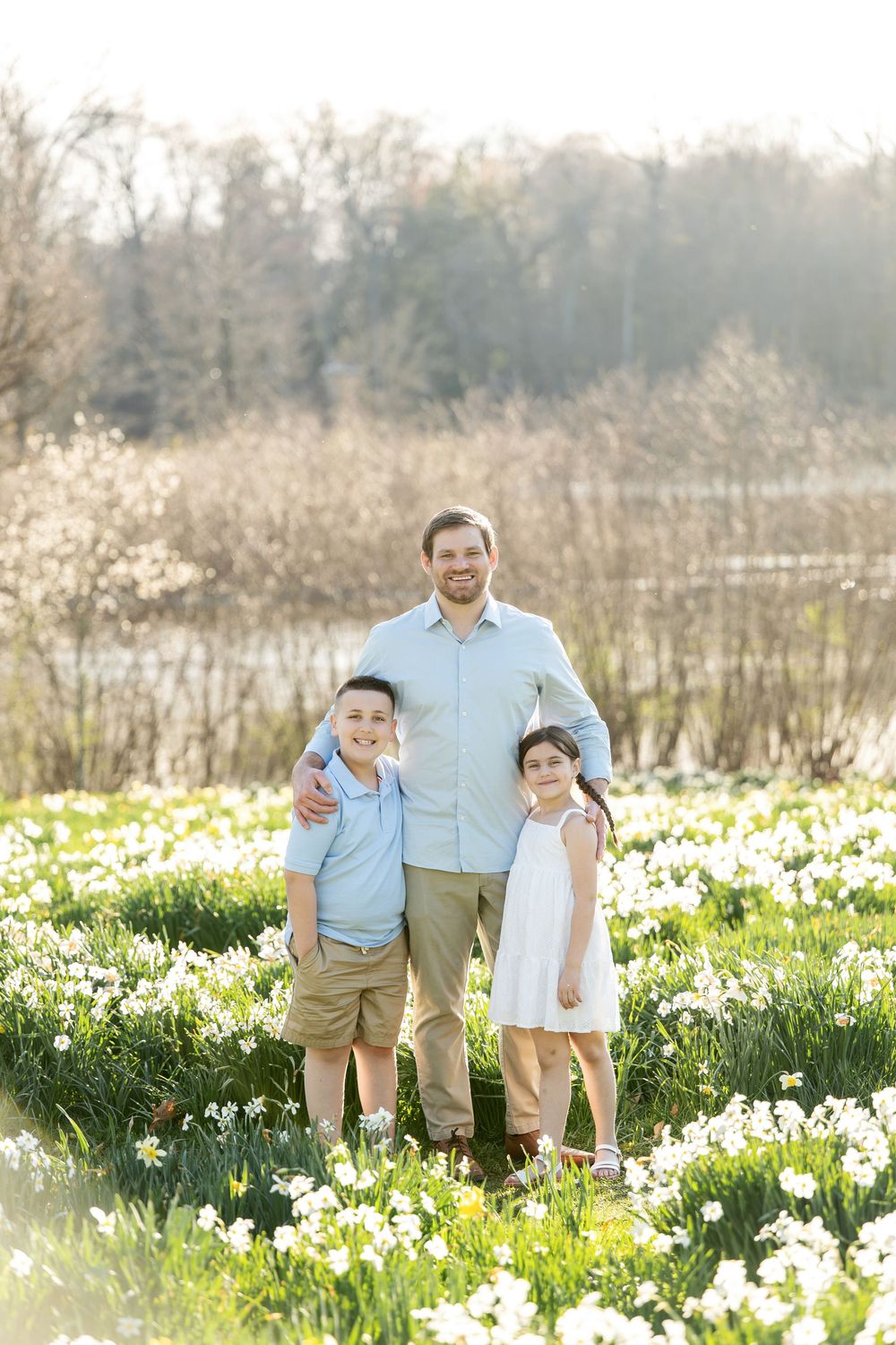 Father smiling with son and daughter in a sunny field of white daffodils during spring.