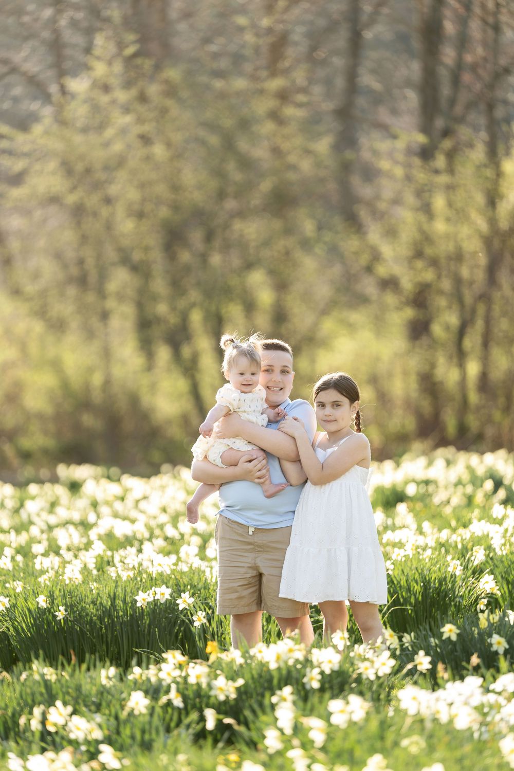 Three siblings posing together in a golden sunlit field filled with white daffodils.