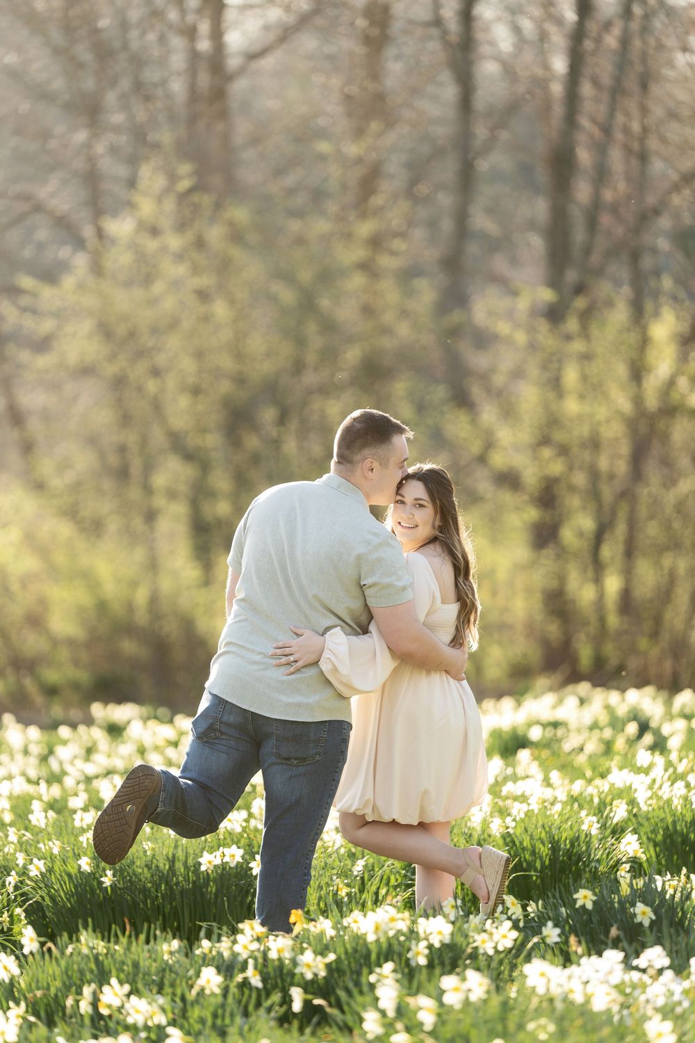 Man lifting woman as she laughs joyfully in a sunny spring daffodil field engagement photo.
