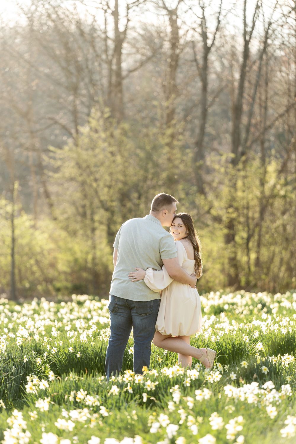 Couple sharing a tender kiss surrounded by white daffodils in a glowing spring outdoor setting.