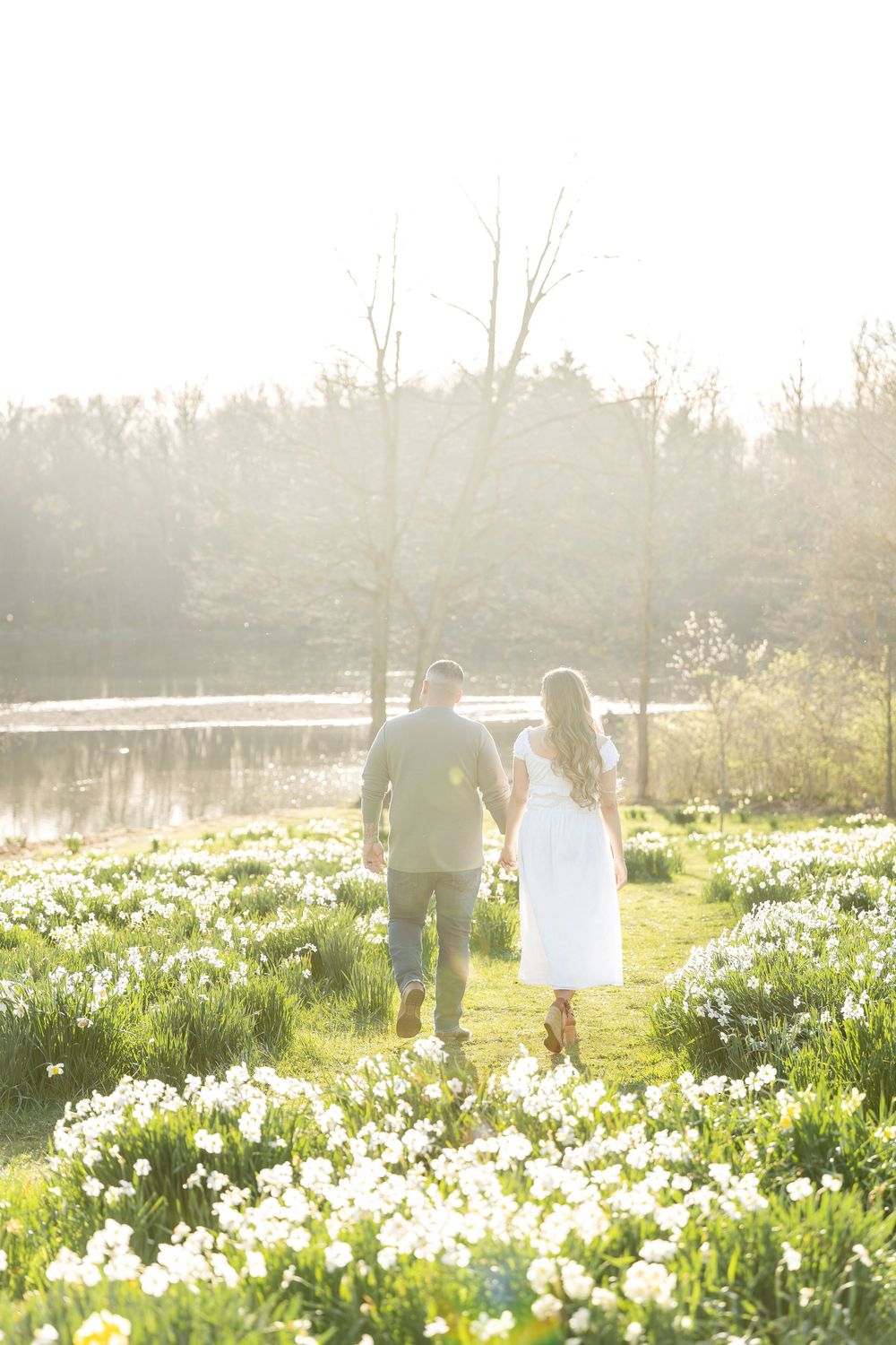 Couple walking hand in hand through a glowing daffodil field toward a sunlit pond in spring.