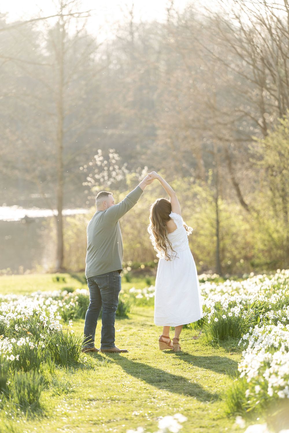 Couple dancing in a sun-drenched daffodil field as man twirls woman in a white dress near a pond.