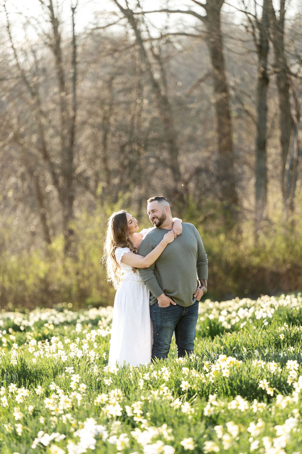 A couple embraces lovingly in a field of white daffodils bathed in warm golden spring sunlight.