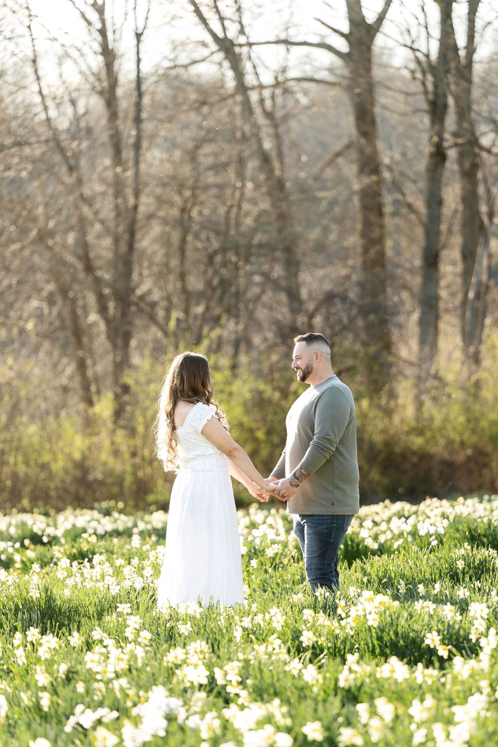 A couple holds hands facing each other in a sunlit daffodil field with bare trees in the background.