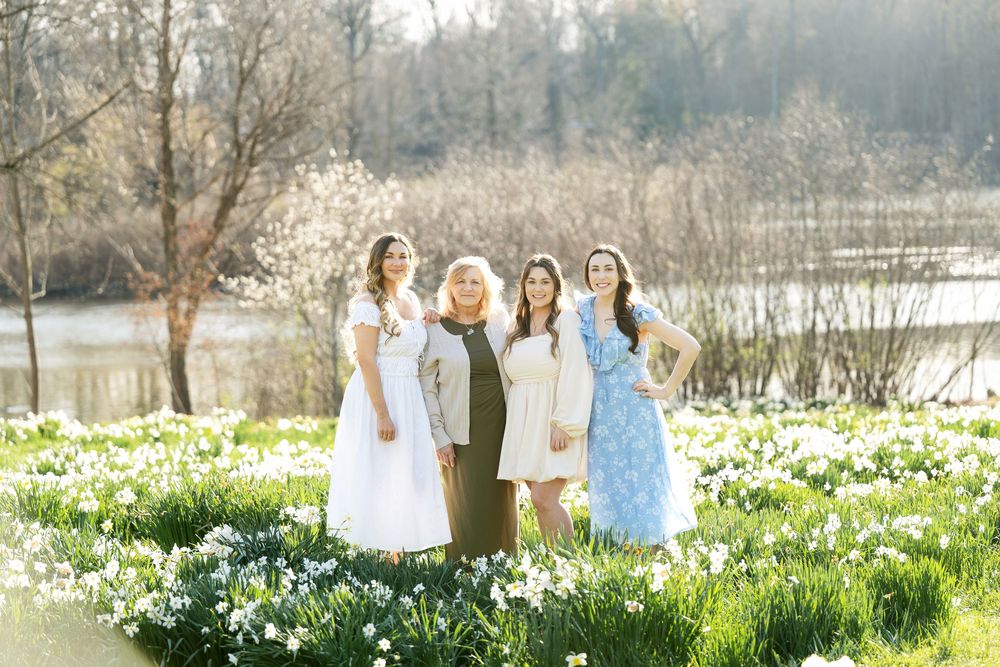 Four women in elegant spring dresses posing together in a lush daffodil field beside a calm pond.