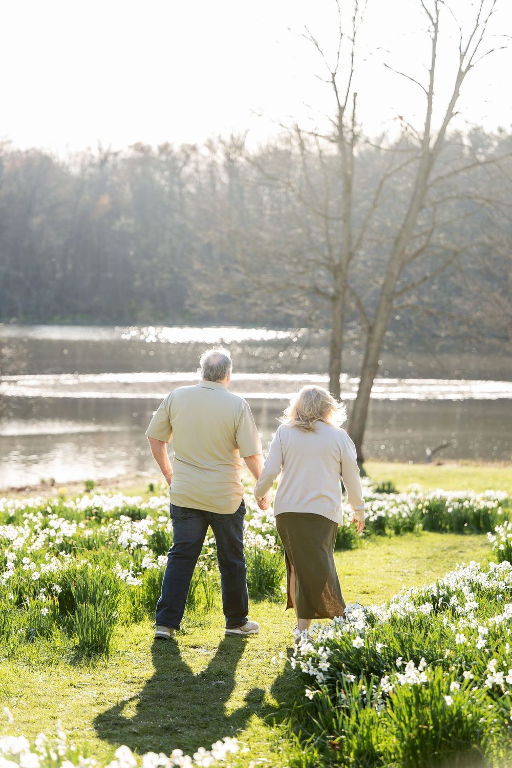 Older couple holding hands walking toward a sunlit pond surrounded by white daffodils in spring.