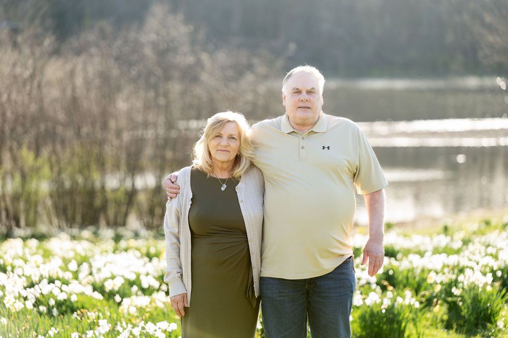 Older couple standing close together in a sunny daffodil field with a peaceful pond in the background.