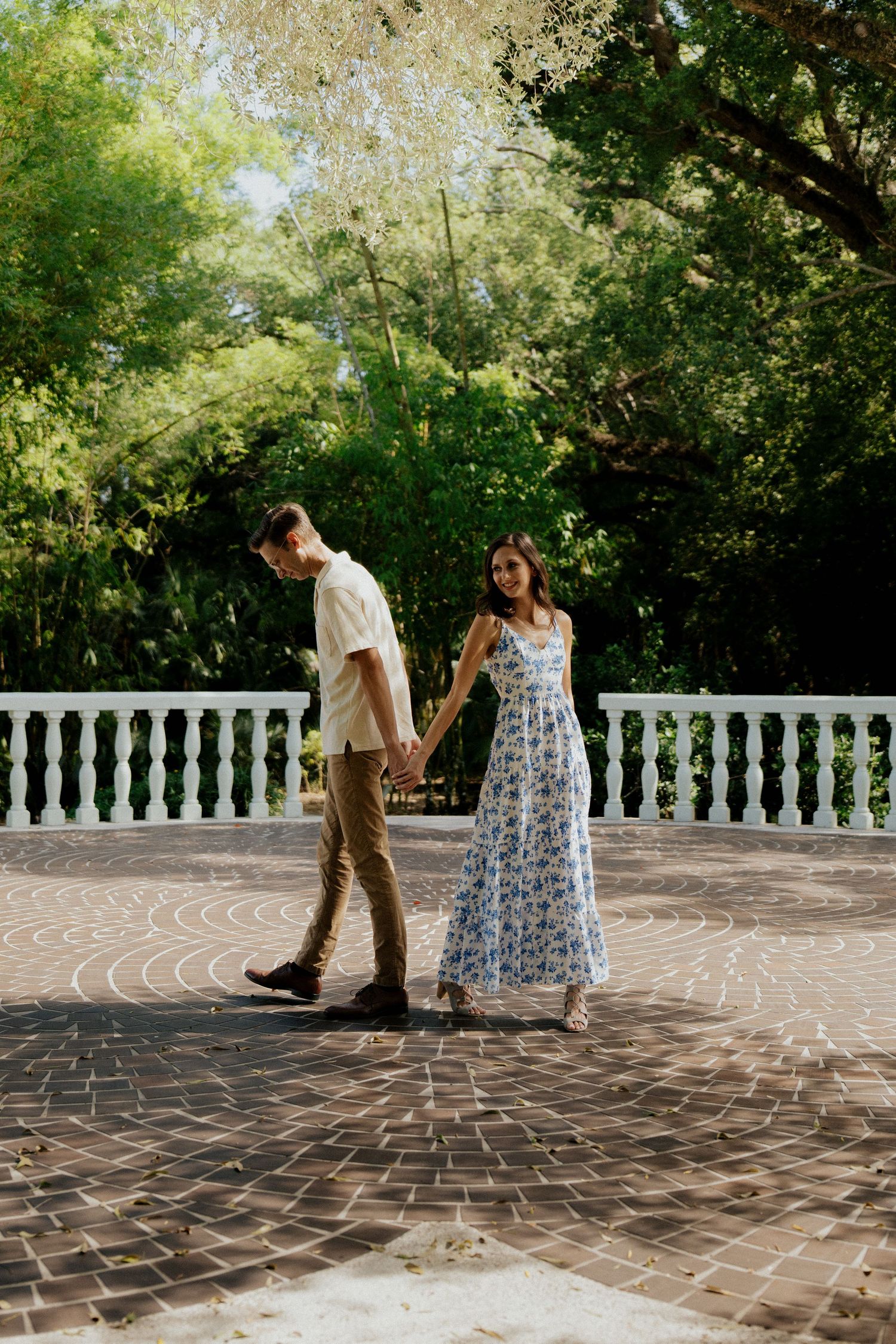 Couple holding hands walking on brick patio surrounded by lush green trees, woman in floral maxi dress smiling.
