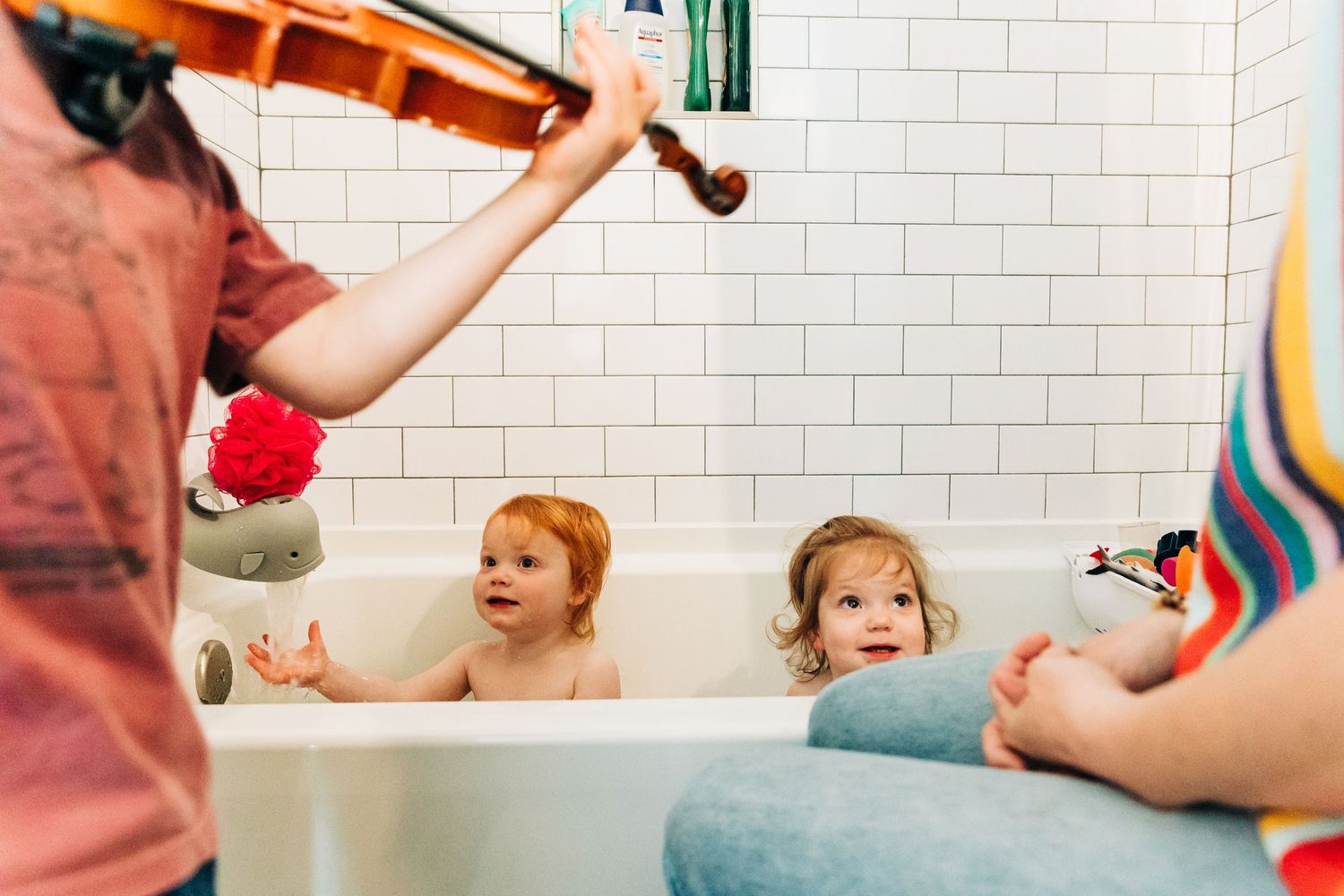 Two toddlers sit in a white tiled bathtub while a sibling plays violin during bath time.