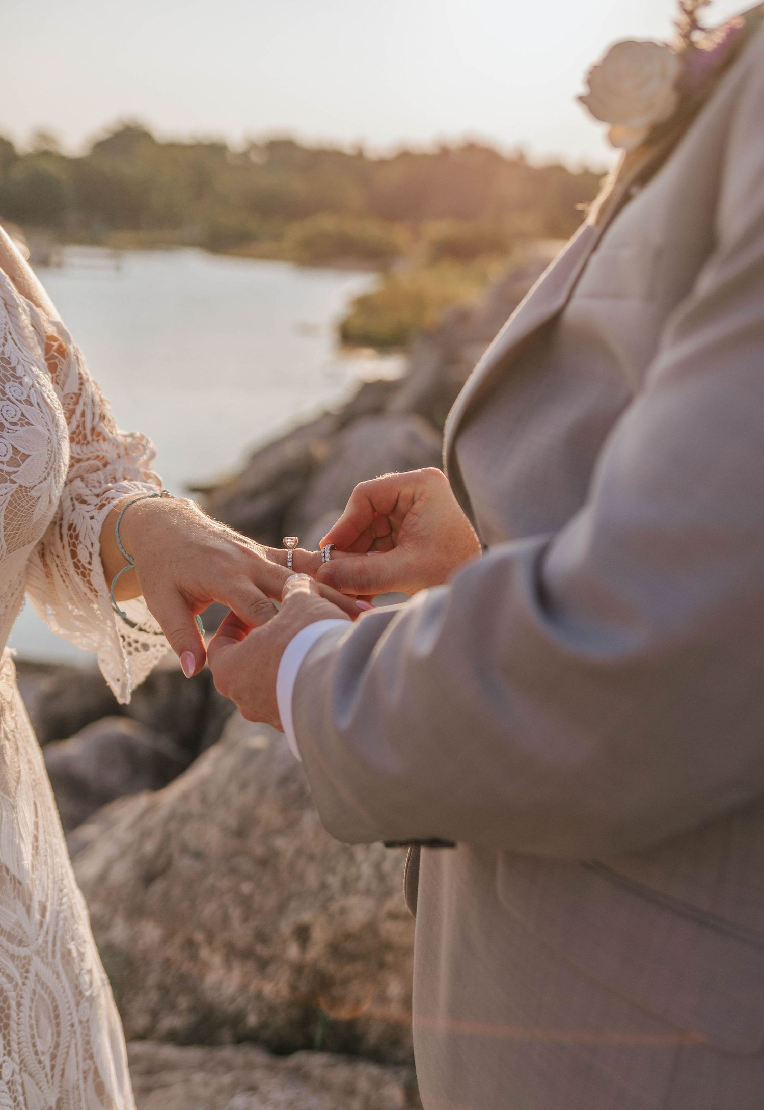 Couple exchanging wedding rings outdoors near rocky waterfront at golden hour sunset ceremony.