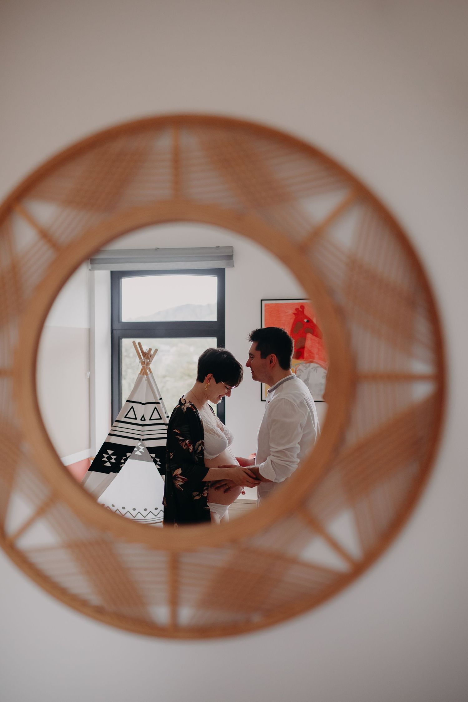 Couple reflected in a round rattan mirror in a childs bedroom with a teepee.