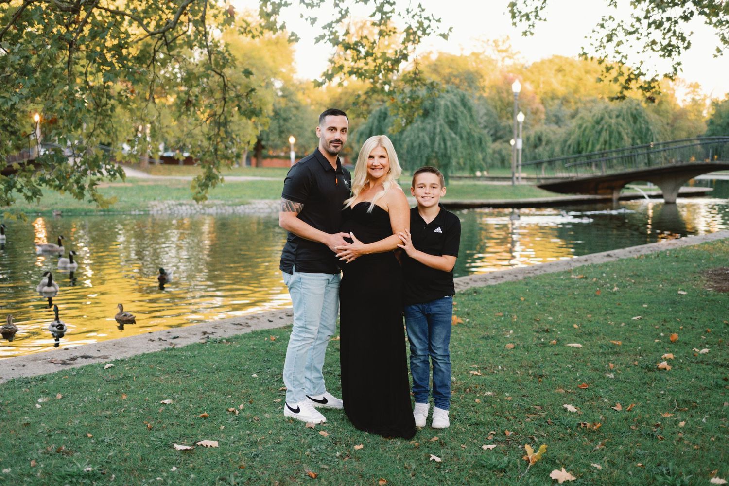 Family of three posing by a serene park pond, pregnant mother in black dress, surrounded by lush green trees.