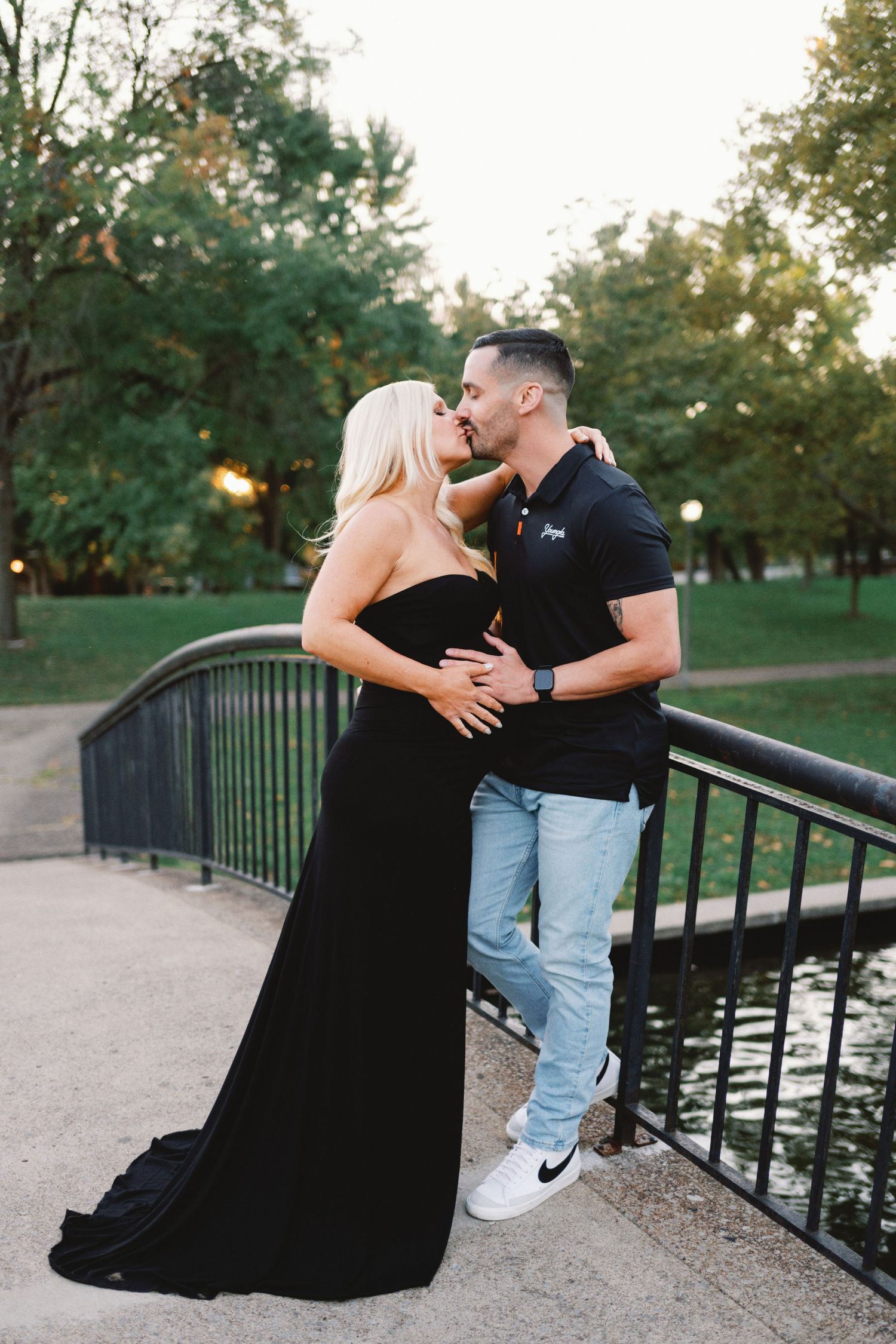Couple sharing a romantic kiss on a bridge in a park, woman in elegant black gown, man in polo shirt and jeans.
