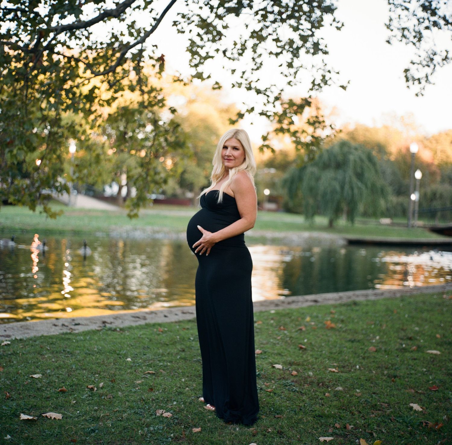 Pregnant woman in black maxi dress posing by a serene park pond at golden hour, cradling her baby bump.