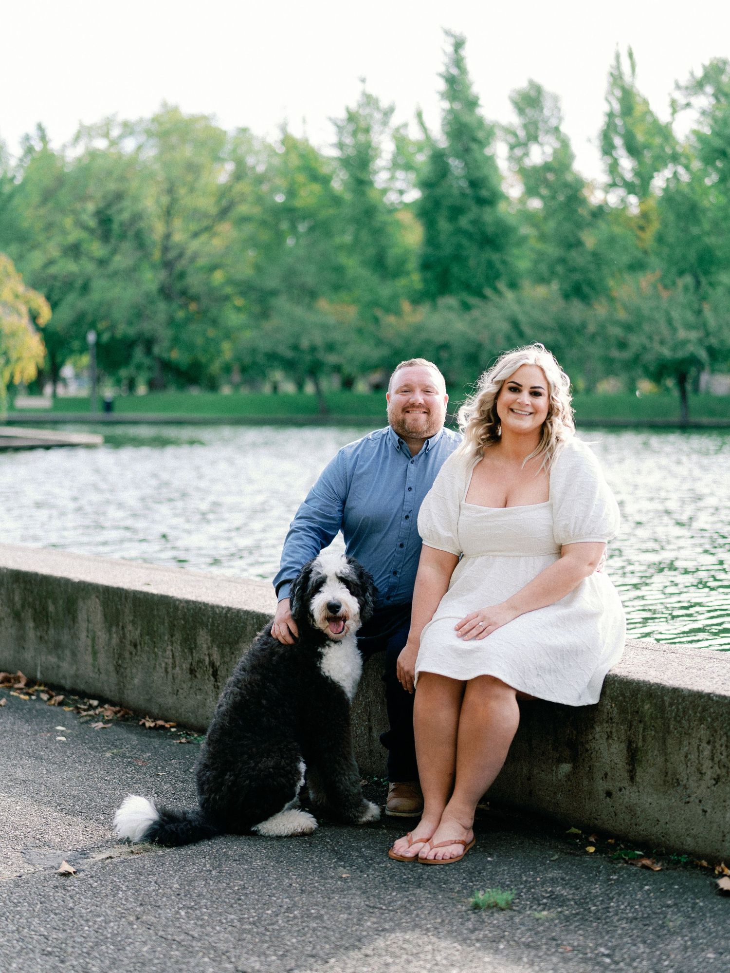 Couple and black and white sheepadoodle dog posing by a serene pond with lush green trees in background.