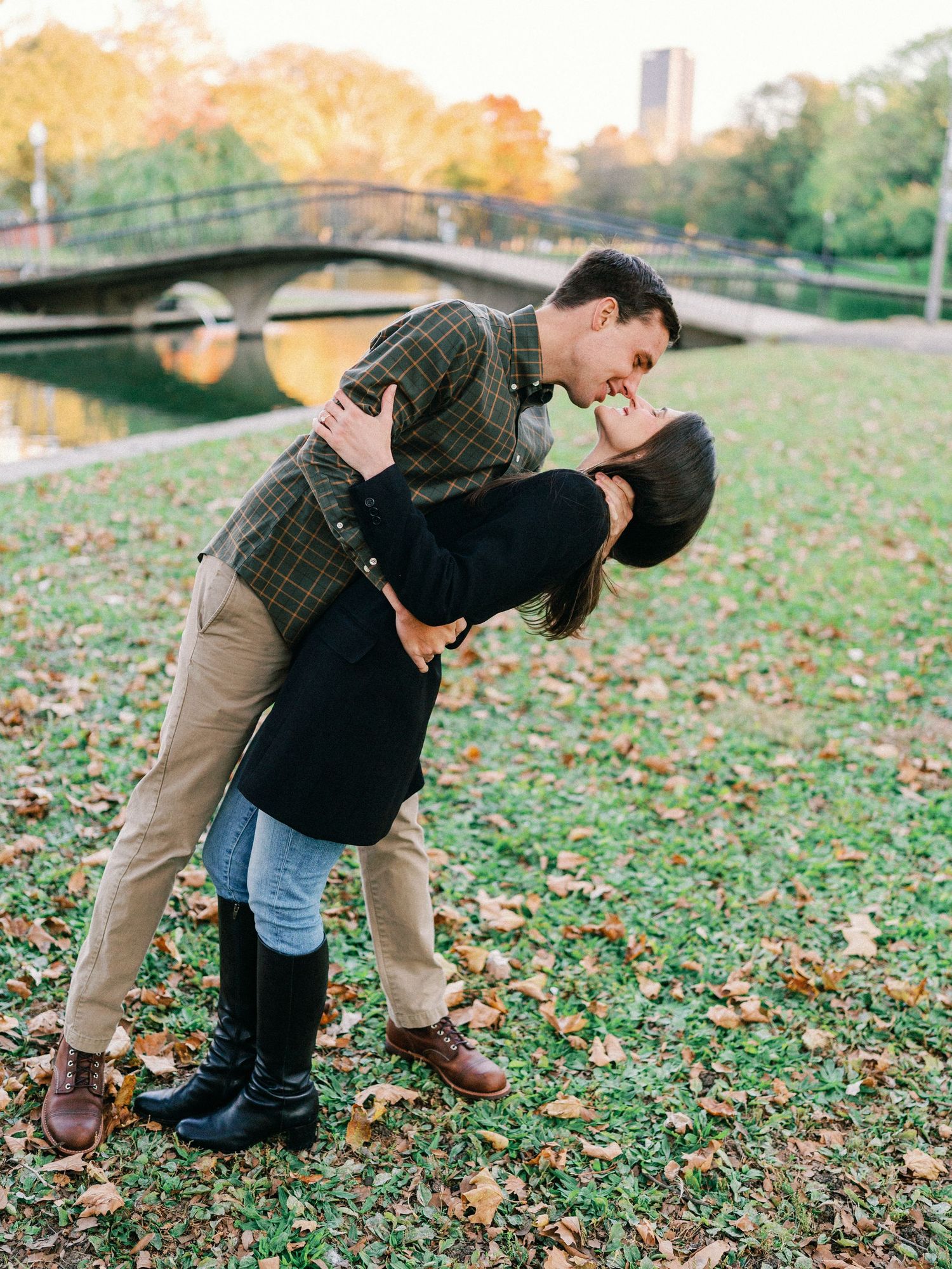 Couple sharing romantic dip kiss in fall park with bridge backdrop.
