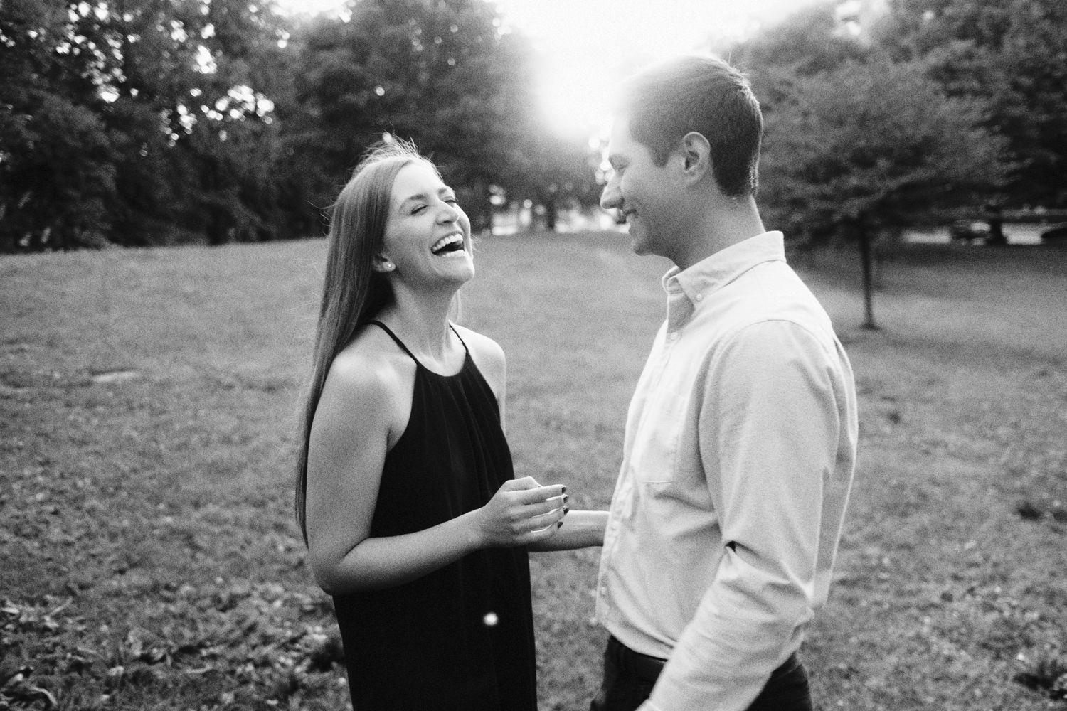 A laughing couple shares a joyful moment outdoors in a black and white photo, woman in dress, man in button-up shirt.