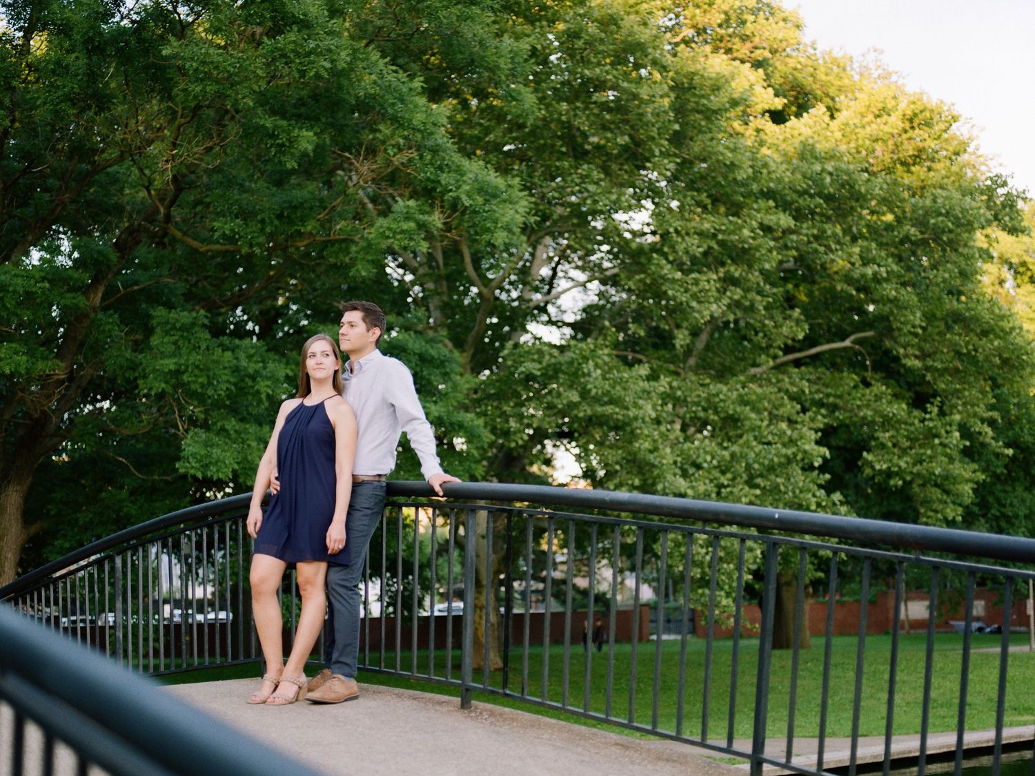 Couple standing on a bridge railing surrounded by lush green trees in a sunny park setting.