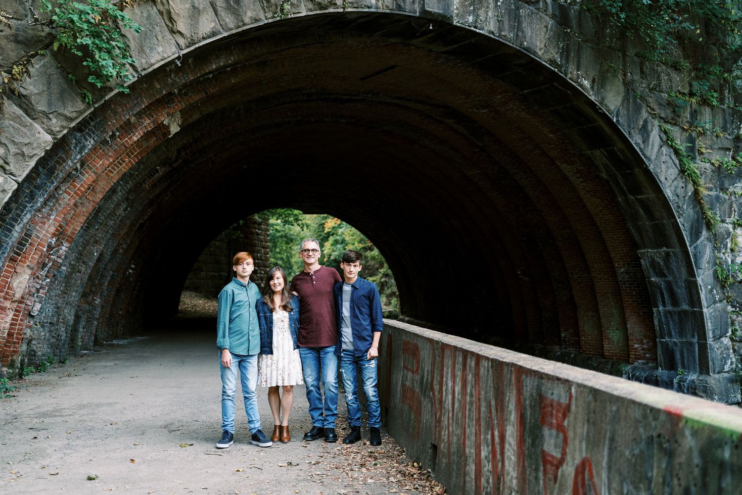 Family of four posing in front of a stone tunnel arch surrounded by lush greenery and graffiti-covered walls.